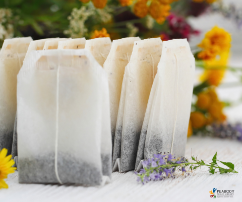 Ten tea bags standing up on a white table with an assortment of flowers in the background and a spring of lavender in the foreground. 