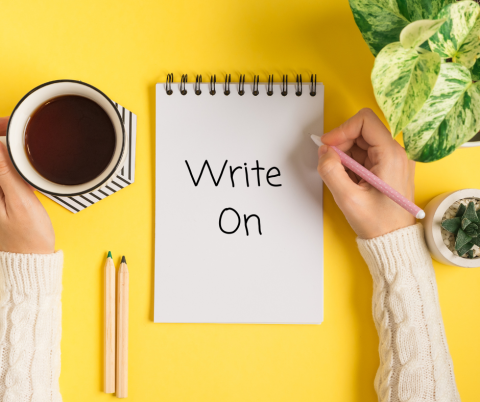 White flip notebook with the words, "Write On" on a yellow background with a hand holding a pen and another hand gripping a coffee mug. Green plant in upper right corner. 