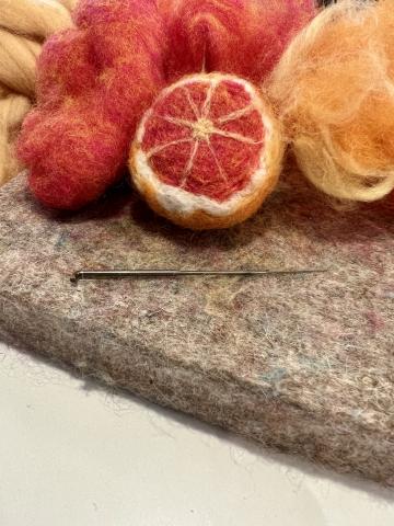 Needle felted grapefruit half on top of felt pad. In front of it is a felting needle. In back of it is orange and yellow wool roving. 